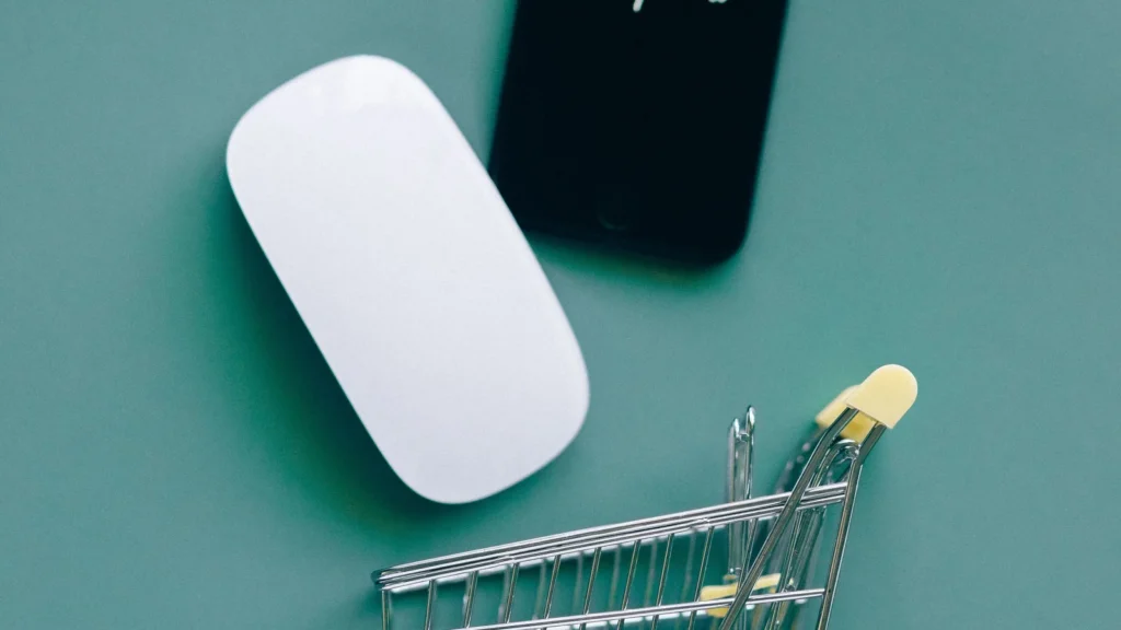 Yeşil bir zemin üzerinde beyaz bir bilgisayar faresi, siyah bir akıllı telefon ve küçük bir metal alışveriş sepeti maketi bulunmaktadır. Görsel, dijital alışveriş ve e-ticaret kavramlarını temsil etmektedir.