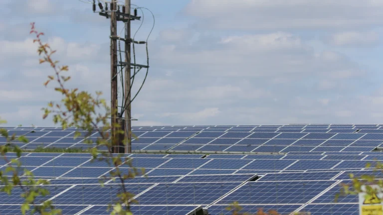 Geniş bir alana yayılmış güneş panelleri (fotovoltaik sistemler) ve ön planda bir elektrik direği ile enerji iletim hattını gösteren dış mekan görseli.