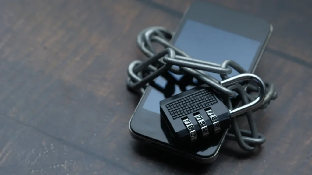 Koyu renkli bir zemin üzerinde, etrafı metal bir zincirle sarılmış ve şifreli bir asma kilit ile kilitlenmiş bir akıllı telefon görseli.