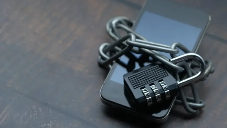 Koyu renkli bir zemin üzerinde, metal bir zincir ve şifreli bir asma kilit ile sarılmış akıllı telefon görseli. Görsel, dijital güvenlik, veri kısıtlaması veya erişim engeli kavramlarını temsil etmektedir.
