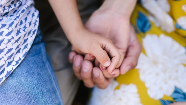 Bir yetişkinin elinin, bir çocuğun elini tuttuğu yakın plan çekim. Görsel, aile bağlarını, ebeveyn-çocuk ilişkisini ve şefkati simgelemektedir.