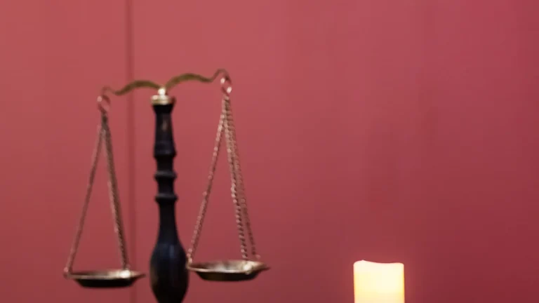Kırmızı bir arka plan önünde duran, adaleti simgeleyen klasik bir terazi ve yanında yanan bir mumun yer aldığı görsel.