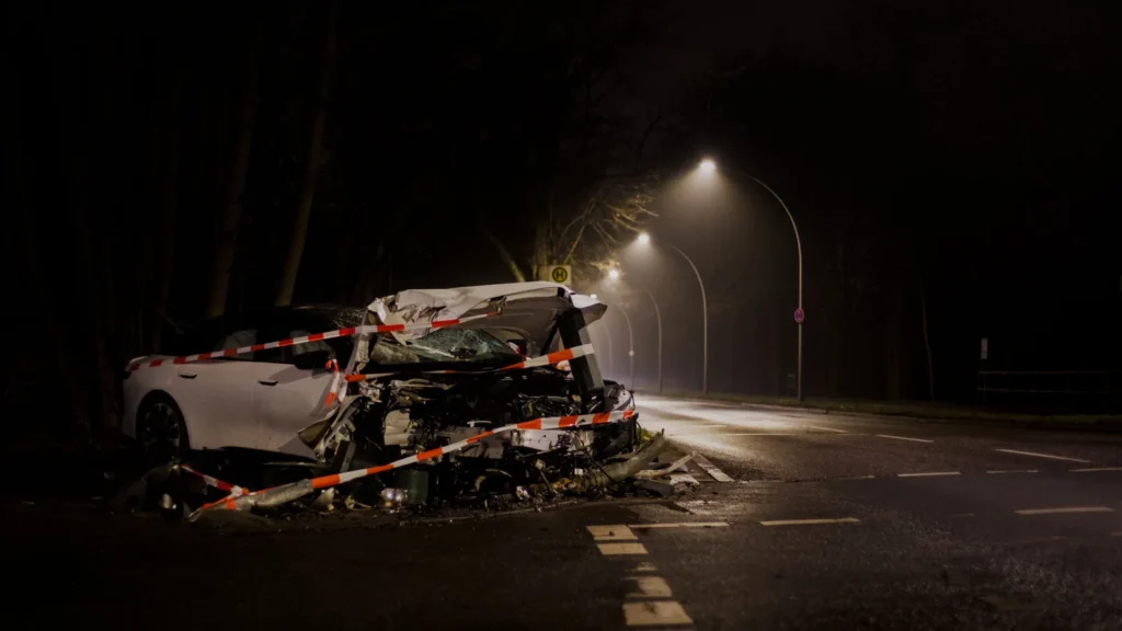 Gece vakti, yol kenarında kaza yapmış, ağır hasarlı ve güvenlik şeridiyle çevrilmiş beyaz bir otomobil.
