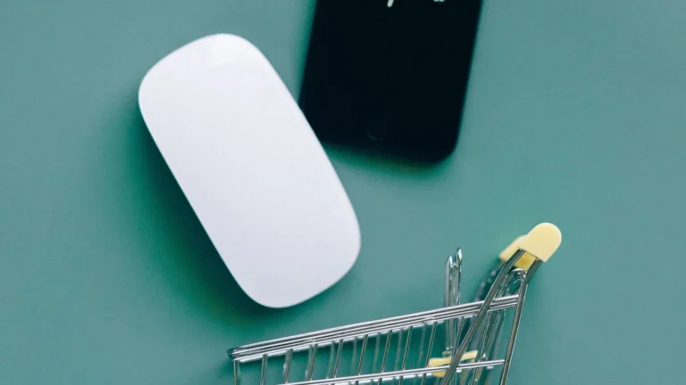 Yeşil bir zemin üzerinde beyaz bir bilgisayar faresi, siyah bir akıllı telefon ve minyatür bir alışveriş sepeti yer almaktadır. Görsel, e-ticaret ve dijital alışveriş süreçlerini temsil etmektedir.