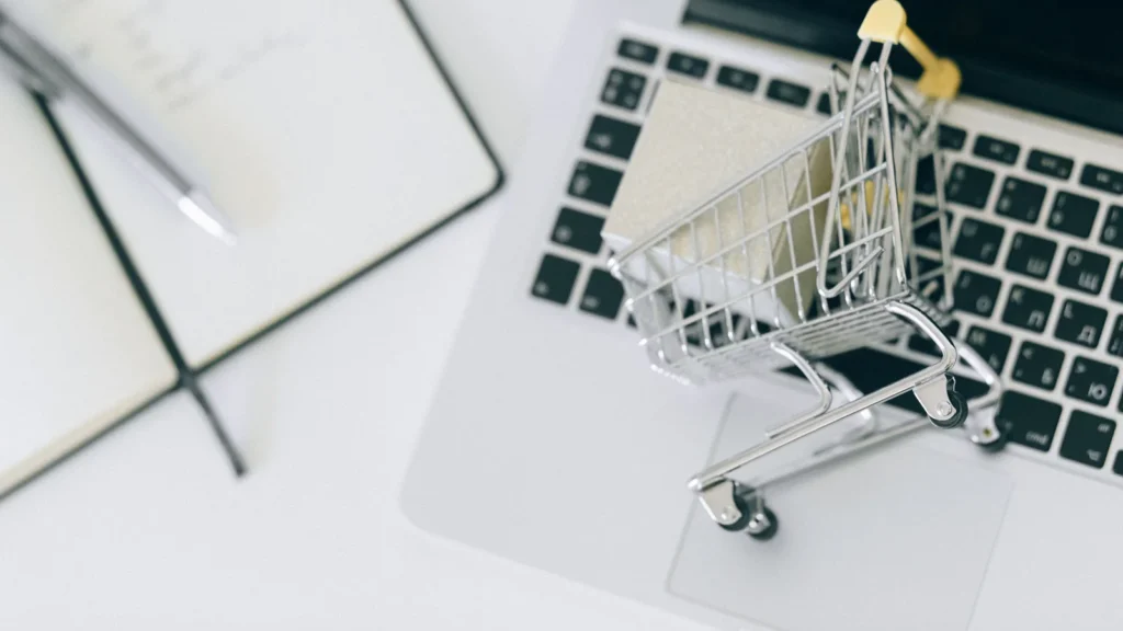 Bir dizüstü bilgisayar klavyesi üzerinde duran minyatür bir alışveriş sepeti ve yanında bir not defteri ile kalem içeren, e-ticaret ve çevrimiçi alışverişi temsil eden görsel.