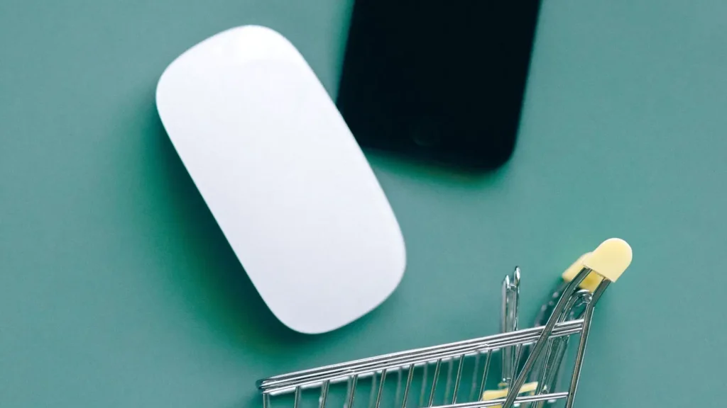 Yeşil bir zemin üzerinde beyaz bir bilgisayar faresi, siyah bir akıllı telefon ve minyatür bir alışveriş sepeti yer almaktadır. Görsel, e-ticaret, çevrimiçi alışveriş ve dijital tüketici deneyimini simgelemektedir.
