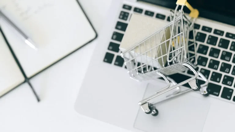 Bir dizüstü bilgisayar klavyesi üzerinde duran minyatür bir alışveriş sepeti ve yanında bir not defteri ile kalem bulunmaktadır. Görsel, e-ticaret, çevrimiçi alışveriş ve tüketici işlemleri kavramlarını temsil etmektedir.