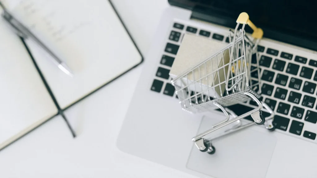 Bir dizüstü bilgisayar klavyesi üzerinde duran minyatür bir alışveriş sepeti, yanında ise not defteri ve kalem bulunmaktadır. E-ticaret ve çevrimiçi alışveriş süreçlerini temsil eden bir görseldir.