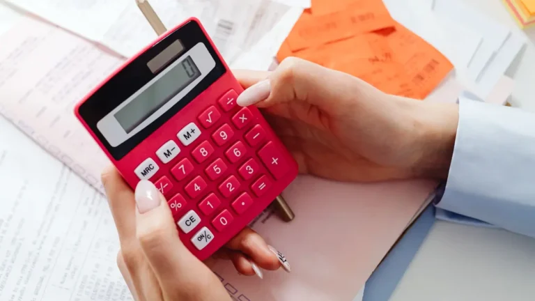 Bir kişinin elinde pembe bir hesap makinesi tuttuğu ve önünde fatura veya fiş benzeri belgelerin bulunduğu yakın plan çekim.