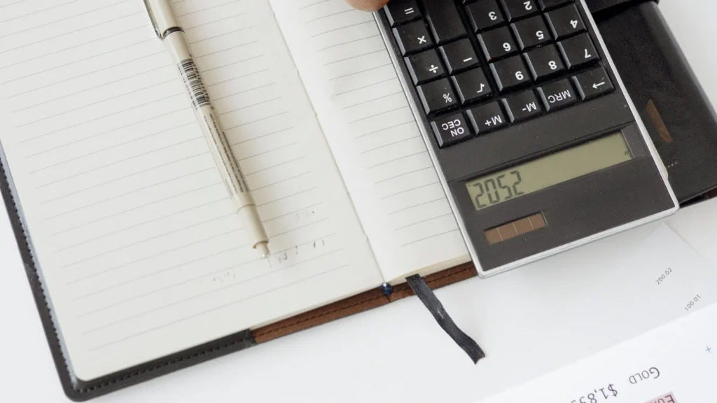 A top-down view of a person using a calculator on an open notebook with a pen, suggesting financial calculation, accounting, or tax-related work.