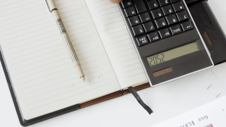 A top-down view of a person using a calculator on an open notebook with a pen, suggesting financial calculation, accounting, or tax-related work.